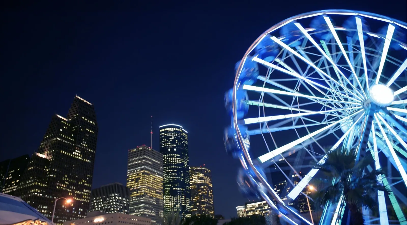 An illuminated Ferris wheel lit blue against the downtown Houston skyline at night